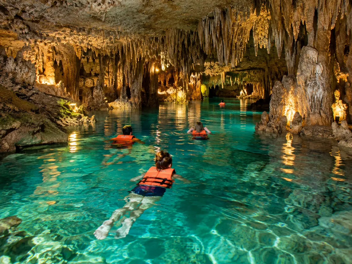 Swimmers in life jackets in a clear underground river among stalactites